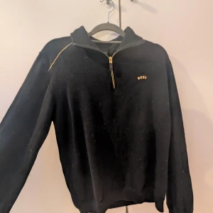 Svart half zip tröja från BOSS - Svart långärmad tröja från BOSS med gulddetaljer och broderad BOSS-logga fram och stort broderi på ryggen. Tröjan har half zip med guldfärgad dragkedja och diskreta sömdetaljer på axlarna. Perfekt för dig som gillar stilren streetwear. Hundhår och inte nopprig. (: skriv för frågor och bilder ⚠️ÄKTA⚠️
