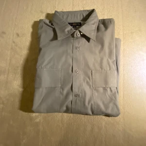 Grå skjorta från The Best, storlek L - Grå skjorta från The Best i storlek L. Klassisk design med två bröstfickor och knappar framtill. Skjortan har en rak passform och är tillverkad i ett mjukt bomullsmaterial som känns skönt mot huden. Perfekt för dig som gillar stilrena och enkla plagg. OBS: Kortärmad!