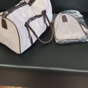 Beige och brun weekendbag i mocka - Snygg och rymlig weekendbag i beige mocka med mörkbruna detaljer och handtag. Väskan har dragkedja upptill och stilren design, perfekt för resor eller övernattningar. Medföljer matchande necessär i samma material och färg.