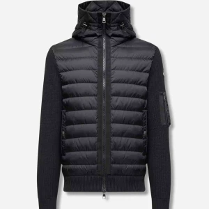 Svart moncler cardigan i storlek L  - Svart dunjacka med stickade ärmar och huva. Jackan har quiltad framsida, dragkedja hela vägen och en ficka med dragkedja på ärmen. Snygg, modern look med ribbade muddar och detaljer i svart.