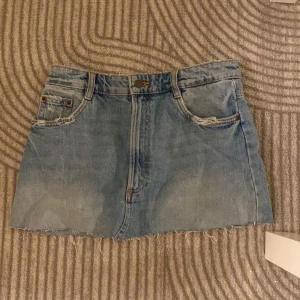 Blå jeanskjol från Zara  - Säljer en blå jeanskjol från Zara i klassisk denim med rå kant och slitna detaljer vid fickorna. Kjolen har fem fickor, bälteshällor och knappstängning framtill. Perfekt för dig som gillar en chill och edgy stil.