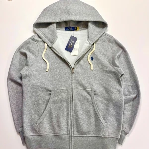 zip up hoodie från Polo Ralph Lauren - Snygg zip up hoodie från Polo Ralph Lauren. skick är 10/10 och aldrig använd. Tillverkad i mjuk bomullsmix, perfekt för chill dagar eller när du vill ha en clean look. Passar dig mellan 165-175. Ny pris runt 2300.