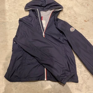 Mörkblå vindjacka från Moncler - Snygg mörkblå vindjacka från Moncler med huva och klassisk logga på ärmen. Jackan har hel dragkedja med rödvit detalj, två fickor med knapp och elastiska muddar. Insidan är grå och materialet är lätt och smidigt, perfekt för blåsiga dagar.