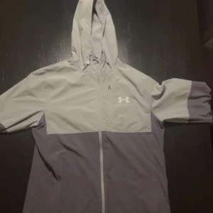 Grå vindjacka från Under Armour - Snygg tvåfärgad vindjacka från Under Armour i ljusgrått och mörkgrått. Jackan har huva, hel dragkedja framtill och lång ärm. Perfekt för blåsiga dagar och sportiga outfits. Under Armour-logga på bröstet ger en sportig vibe.