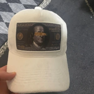 Trucker keps - Säljer en vit trucker keps från D’Arigato med mesh på sidorna och ett stort patch framtill med dollartryck i svart och guld. Skön passform och cool detalj som sticker ut. Perfekt för dig som gillar streetwear och vill ha något unikt.