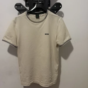 Vit BOSS t-shirt med svart logga - Snygg vit t-shirt från BOSS med svart logotyp på bröstet. T-shirten har korta ärmar och ribbade muddar med svarta ränder vid hals och ärmslut. Tillverkad i mjuk bomull för skön känsla. Perfekt för dig som gillar stilrena och klassiska plagg.