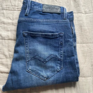 Blå Replay jeans med snygg tvätt - Säljer ett par blå jeans från Replay med klassisk femficksdesign och snygga slitningar. Jeansen har en cool tvätt och tydliga sömmar på bakfickan. Perfekta för dig som gillar en avslappnad men stilren look.