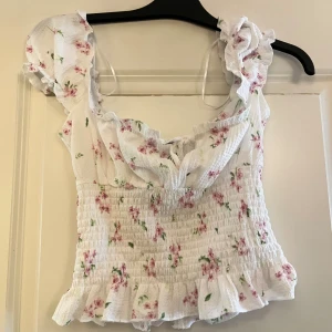 Blommig vit blus från Gina Tricot - Supersöt blommig blus från Gina Tricot i vit med rosa och gröna detaljer. Blusen har volangkant, smockad midja, knyt framtill och puffiga axlar. Materialet är mjukt och lätt, perfekt för varma dagar. Säljer då den inte kommer till användning!
