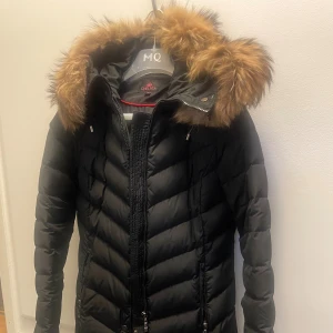 Svart lång dunjacka med fuskpäls - Svart lång dunjacka från Chelsea med snyggt quiltat mönster och huva med fluffig brun fuskpäls. Jackan har dragkedja framtill, två fickor med dragkedja och en figurnära passform. Perfekt för kalla vinterdagar. Priset går att diskutera.