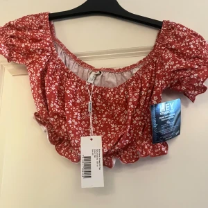 Röd blommig volangblus från Nelly - Superfin röd blus från Nelly med vita blommor och volangdetaljer. Modellen har puffiga korta ärmar och en söt, rynkad kant vid midjan. Perfekt för dig som gillar romantiska och trendiga toppar med offshoulder-vibe.