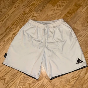Vita Adidas Cimalite shorts med snörning - Säljer ett par vita Adidas Cimalite shorts i storlek M. De har elastisk midja med snörning, svart Adidas-logga på benet och svart kant längst ner. Materialet är lätt och andas, perfekt för sport eller chill. Klassisk loose fit och stilren design.