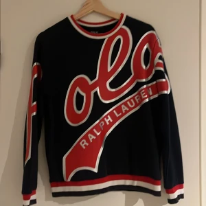 Svart långärmad tröja Polo Ralph Lauren - Tja säljer en Cool marinblå  långärmad tröja från Polo Ralph Lauren med stor röd och vit logga framtill. Tröjan har ribbade muddar och krage i rött och vitt, samt breda ränder vid ärmslut och nederkant. Pris kan diskuteras vid snabb affär! 