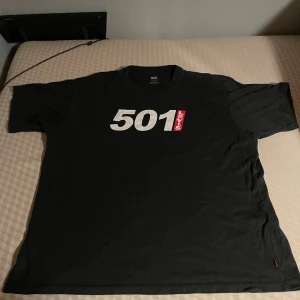 Svart Levi's 501 t-shirt - Svart t-shirt från Levi's med stort vitt 501-tryck och röd Levi's-logga på bröstet. Klassisk rund hals och korta ärmar. Tillverkad i mjuk bomull som är skön mot huden. Perfekt för dig som gillar streetwear och ikoniska tryck.