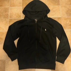Svart tracksuit från Polo Ralph Lauren - Svart tracksuit från Polo Ralph Lauren med klassisk vit broderad logga på bröstet. Tröjan har huva med snörning, dragkedja framtill och två fickor. Materialet är mjuk bomull och passformen är normal. Perfekt för chill dagar och streetwear.     Oanvänd! 