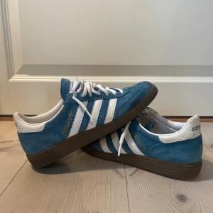 Adidas Spezial blå sneakers mocka - Adidas Spezial sneakers i blå mocka med vita detaljer och klassiska tre ränder på sidan. Skorna har vit snörning, brun gummisula och rund tå. Adidas-logga på hälen och gul Spezial-tryck på sidan. Perfekt för dig som gillar retrostil och streetwear. Använda bokstavligen ett fåtal gånger, skick 9/10