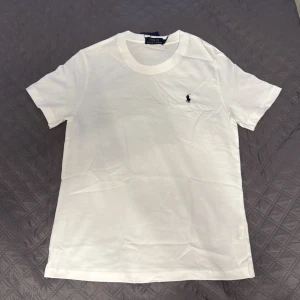 Vit t-shirt från Polo Ralph Lauren - Vit t-shirt från Polo Ralph Lauren med klassisk svart broderad logga på bröstet. Modellen är kortärmad och har en rund halsringning. Tillverkad i mjuk bomull för skön komfort och stilren look.