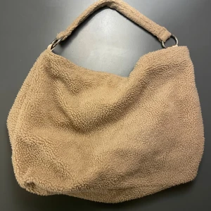 Stor beige axelväska i teddymaterial - Väldigt praktisk och mysig väska som passar perfekt för att bära flera saker! Den har en magnetisk knapp som gör den enkel att stänga ❤️ på insidan har den ett lent material och en liten ficka på sidan! 