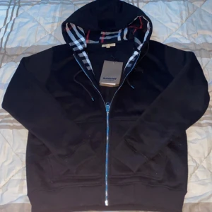 Svart hoodie från Burberry - Säljer en stilren svart hoodie från Burberry med dragkedja och klassiskt rutigt mönster i luvan. Tröjan har långa ärmar och en broderad logga på bröstet. Perfekt för en avslappnad och trendig look.