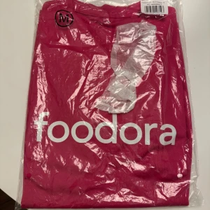 Foodora rosa t-shirt i polyester - Säljer en rosa t-shirt från Foodora med vit logga tryckt på bröstet, ryggen och ärmen. T-shirten har korta ärmar och är gjord i ett lätt och ventilerande polyestermaterial. På baksidan finns coola vita ikoner med cykel, mobil och burgare. Tröjan är i storlek M och är helt ny från paketering! Svarar snabbt!😇