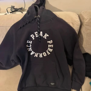 Marinblå hoodie från Peak Performance - Säljer en marinblå hoodie från Peak Performance med stor vit logga i cirkel på bröstet. Tröjan har huva med snörning och en klassisk magficka. Perfekt för dig som gillar sportig och avslappnad stil.