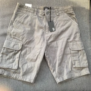 Grå cargo shorts från 157, storlek M - Ett par gråa shorts med fickor på sidorna. Shortsen har en knappstängning och en svart etikett med texten '157.' Beninnerlängd: 25 cm ﻿Midja: 88 cm ﻿100% bomull ﻿Knapp och dragkedja ﻿5 ficks modell  Inga bilder när den sitter på mig! Katter finns i hemmet!