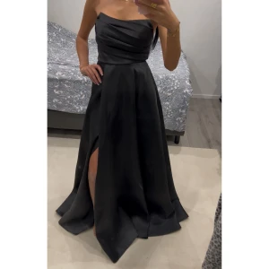 Svart axelbandslös långklänning - Elegant svart axelbandslös långklänning med draperad överdel och snygg slits framtill. Klänningen har en klassisk och stilren siluett som ger en lyxig känsla. Perfekt för dig som vill sticka ut med en sofistikerad look. Använd endast en gång. Storleken går att justera bak med band så passar 34-38