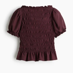 Vinröd blus med puffärm och volang - Supersöt vinröd blus med puffärmar och smockad kropp. Toppen har volangkant nedtill och vid ärmslut. Modellen är figurnära med korta ärmar och rundad hals. Helt ny, nypris var 250💗