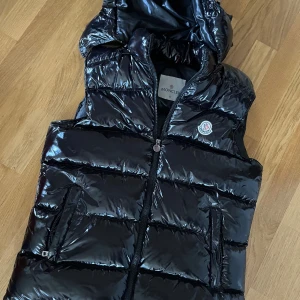 Svart dunväst från Moncler med huva - Snygg svart dunväst från Moncler i glansigt material med huva och dragkedja framtill. Västen har två fickor med dragkedja och ett broderat Moncler-märke på bröstet. Perfekt för lager-på-lager och streetstyle. PM för bilder/frågor