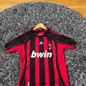 AC Milan Kaka Adidas matchtröja - Säljer en klassisk AC Milan replika fotbollströja med Kaka och nummer 22 på ryggen. Adidas, officiell matchtröja med Bwin sponsortryck, klubbmärke och Champions League-patch på ärmen. Röd och svart med guldtryck, korta ärmar och randigt mönster. Perfekt för fotbollsfantaster eller samlare. Skick 9,5/10 Hör av er vid frågor! Pris kan diskuteras