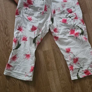 Vita blommiga croppade byxor - Säljer ett par vita croppade byxor med rosa och grönt blommönster. Byxorna har raka ben, två bakfickor och är tillverkade i ett mjukt bomullsmaterial. Perfekt för dig som gillar färgglada och unika plagg med somrig vibe.