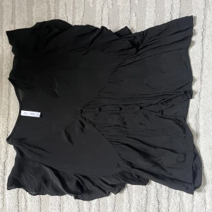 Svart fin blus från MNG - Svart blus från MNG med v-ringad hals och dekorativa knappar framtill. Blusen har en flowy siluett och är tillverkad i ett mjukt, glansigt material som ger en elegant känsla. Perfekt för dig som gillar stilrena och enkla plagg med snygga detaljer.