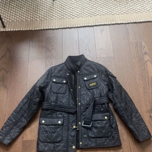 Svart quiltad jacka Barbour junior XXL motsvarar stl 14-15 år - Quiltad svart jacka från Barbour International med bälte i midjan, guldiga knappar och dragkedja. Jackan har flera fickor framtill, hög krage och klassisk Barbour-logga på bröstet. Perfekt för dig som gillar stilren och funktionell design. Storleken är junior XXL vilket motsvarar stl 14-15 år! Skickar oftast inom 24h📦