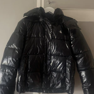 Svart pufferjacka Calvin Klein Jeans S - Snygg svart pufferjacka från Calvin Klein Jeans med glansig finish och diskret CK-logga på bröstet. Jackan har huva, dragkedja och tryckknappar framtill samt stora fickor. Perfekt för kalla dagar.💞