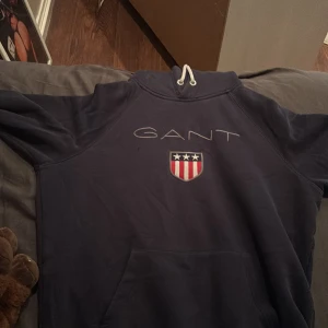 Marinblå hoodie från GANT - Snygg marinblå hoodie från GANT med stor broderad logga och emblem på bröstet. Tröjan har huva med vita snören och en klassisk känguruficka framtill. Perfekt för chill dagar och har en mjuk insida som gör den extra skön.