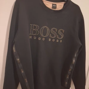 Svart sweatshirt från Hugo Boss - Svart sweatshirt från Hugo Boss med stort guldfärgat logotryck på bröstet och dekorativa band med BOSS-tryck längs ärmarna. Klassisk rund halsringning och långa ärmar. Perfekt för en stilren och trendig look.