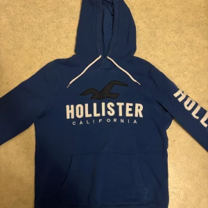 Blå hoodie från Hollister med tryck - Blå hoodie från Hollister med stor logga och fågel på bröstet samt text på ena ärmen. Tröjan har huva med vita snören och en stor magficka. Mjuk insida och klassisk passform, perfekt för chill dagar. En liten defekt på bild 5
