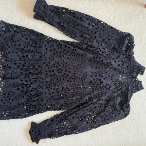 Svart broderad kortklänning med spets - Supersnygg svart kortklänning med långärmade ärmar och broderade spetsdetaljer. Klänningen har ett unikt hålmönster med blommiga inslag och volang vid nederkanten. Tillverkad i bomull, perfekt för dig som gillar statement-plagg med coola detaljer.