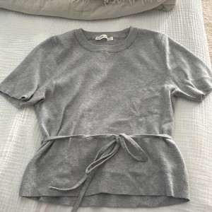 Grå stickad topp med knyt från Pull&Bear - Säljer en grå stickad topp från Pull&Bear i storlek L. Toppen har korta ärmar, rund halsringning och ett snyggt knytband i midjan som ger en fin siluett. Perfekt för dig som gillar enkel men stilren design.