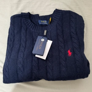 Mörkblå stickad tröja Polo Ralph Lauren - Kabelstickad mörkblå tröja från Polo Ralph Lauren med rund halsringning och klassisk röd broderad logga på bröstet. Tröjan är i mjuk bomull och har ribbade muddar vid ärmslut och nederkant. Perfekt för en stilren och tidlös look.                                                                                       Pris går att diskutera
