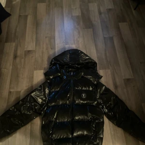 Svart Trapstar pufferjacka med huva - Säljer en svart, glansig pufferjacka från Trapstar med stor huva och dragkedja framtill. Jackan har broderad Trapstar-logga på ryggen och rund logotyp på bröstet. Perfekt för kalla dagar och riktigt snygg streetstil.