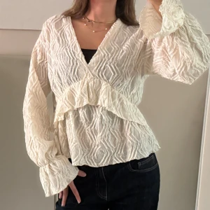 Vit omlottblus med volangdetalj - Säljer en superfin vit blus med omlottkänsla och v-ringning. Blusen har långärmade, lätt puffiga ärmar. Perfekt att styla med jeans eller kjol. Den är i en ljusgul/crémevit färg. Använd 2 gånger och är i utmärkt skick!