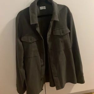 Mörkgrön overshirt från These Glory Days - Snygg mörkgrön overshirt från These Glory Days i storlek medium. Jackan har klassisk krage, två bröstfickor med knappar och raka ärmar. Perfekt lager-på-lager-plagg med stilren look och mjukt material. Nyss köpt aldrig använd för stor mig mig