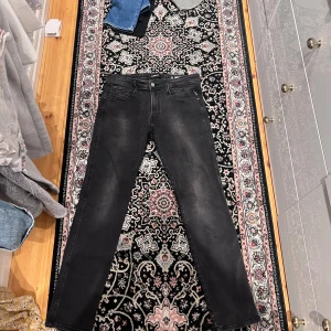 Svarta Replay Anbass jeans, stl 33/34 - Svarta Replay Anbass jeans i klassisk femficksmodell. Jeansen har en snygg tvättad finish och raka ben. Märkesdetaljer på fickorna och bak, samt diskret Replay-logga. Perfekta för dig som gillar en stilren och modern look.