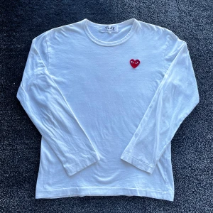 CDG Långärmad tröja  - Säljer en vit långärmad tröja från Comme des Garçons PLAY med det ikoniska röda hjärtat broderat på bröstet. Tröjan är i mjuk bomull och har en klassisk rund hals. Perfekt för dig som gillar stilrena och trendiga plagg. Skick 8/10, finns ett litet hål men inget som syns, storlek L men passar även M, äkta. Pris går att diskutera✅