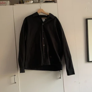 Svart overshirt från Carhartt - Svart overshirt från Carhartt med klassisk krage och stora knappar framtill. Skjortan har två diskreta fickor framtill och är tillverkad i ett slitstarkt bomullstyg. Långärmad modell med enkel, clean look som passar till det mesta.