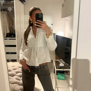 Vit blus med volang och knappar - Supersöt vit blus i bomull med volangdetaljer och knappar framtill. Lätt v-ringad med rynkade partier och lång ärm. Perfekt för dig som gillar en romantisk och chill vibe. Sitter lite loose och har en flowy känsla.