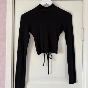 Svart croppad polotopp med snörning - Säljer en svart croppad polotopp med långa ärmar. Toppen har öppen rygg som ger en snygg och trendig look med en snörning. Perfekt för dig som gillar stilrena plagg med en twist.