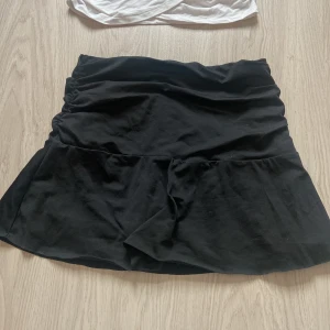 Svart volangkjol i bomull - Supersöt svart kortkjol med volangkant och lätt rynk vid midjan. Kjolen är gjord i mjuk bomull och har en bekväm passform som sitter snyggt på höften. Perfekt för dig som gillar en enkel men trendig look.