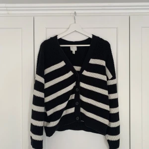 Randig svartvit kofta från H&M - Säljer en stickad kofta från H&M i svart och vitt med breda, diagonala ränder. Koftan har v-ringning, stora knappar framtill och långa ärmar. 