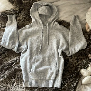 Grå stickad hoodie från Garage - Mysig grå stickad hoodie från Garage med huva och snörning. Tröjan har ribbade muddar vid ärmslut och nederkant samt en praktisk magficka. Perfekt för chill dagar och har en loose passform som ger en avslappnad vibe.